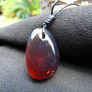 Puede incluir: Un collar con colgante pulido en forma de lágrima. La piedra de color ámbar pasa de un rojo intenso en la base a un azul oscuro en la parte superior. El colgante está suspendido de un cordón negro, anudado en la parte superior.
