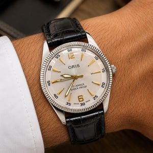 Puede incluir: Un reloj de pulsera Oris con esfera plateada, manecillas e índices dorados, y números dorados en las 6 y las 12. El reloj cuenta con un bisel estriado, una correa de cuero negro y el texto Oris, 17 Jewels, Shock Proof, y Swiss Made en la esfera.