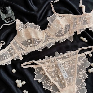 Puede incluir: Un conjunto de lencería color melocotón claro con un diseño de encaje floral. El conjunto incluye un sujetador y un tanga, ambos adornados con delicadas perlas. El sujetador tiene tirantes ajustables y el tanga tiene una etiqueta con la talla M. El conjunto se muestra sobre tela de satén negro.