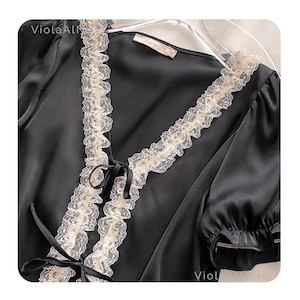 May include: Black satin top with cream-colored lace trim along the neckline and sleeves. The top features a V-neck design with a bow detail at the center. The sleeves are short and have a ruffled edge. The top is hanging on a hanger.