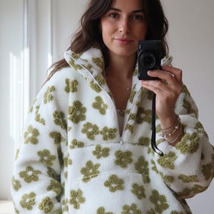 Op de afbeelding: Witte fleece trui met olijfgroen bloemenpatroon. De jas heeft een halve ritssluiting en een hoge kraag. De zachte, getextureerde stof is bedekt met herhaalde bloemvormen. Een gezellige en stijlvolle buitenkleding.