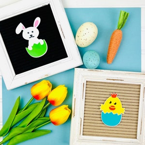 May include: Two white picture frames with black felt boards inside. One frame has a white bunny in a green egg, the other has a yellow chick in a blue egg. A bouquet of yellow tulips and a carrot are on a blue background.