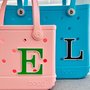 May include: Two colorful tote bags. One is pink with a green letter "E" and red polka dots. The other is blue with a black letter "L". Both bags have handles and the brand name "bogg" on the bottom.