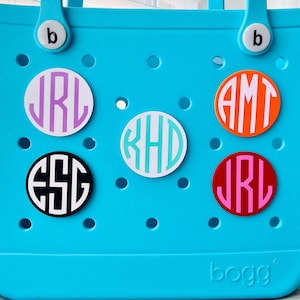 May include: A turquoise tote bag with several round monogrammed decals in various colors. The decals feature initials in different fonts and colors, including purple, orange, red, black, and teal. The bag has a handle and the brand name "bogg" is visible.