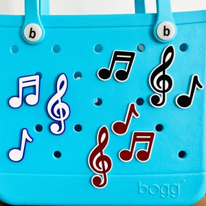May include: A turquoise tote bag adorned with various music note charms in black, red, and white. The bag has two handles and the brand name "bogg" is visible on the bottom right. The charms are in different sizes and styles.