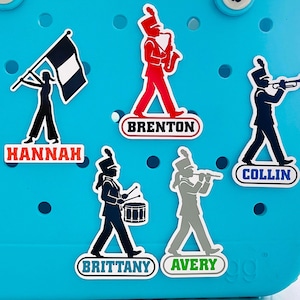 May include: Five decorative shoe charms attached to a bright blue perforated surface. The charms feature silhouettes of marching band members in various colors, each with a name tag: Hannah, Brenton, Collin, Brittany, and Avery.
