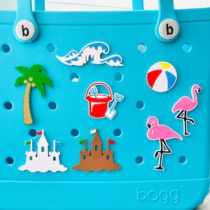 K&ouml;nnte beinhalten: Eine t&uuml;rkisfarbene Tragetasche mit Strandmotiven. Die Motive umfassen eine Palme, Wellen, einen Strandball, einen Sandeimer, eine Schaufel, zwei Flamingos und zwei Sandburgen. Auf der Tasche steht das Wort "bogg".