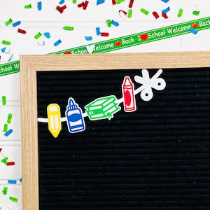 May include: A black letter board with a white string of school supply cutouts. The cutouts include a pencil, glue bottle, stack of books, crayon, and scissors. The letter board is surrounded by a wooden frame. The background is white with confetti.