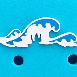 K&ouml;nnte beinhalten: Ein wei&szlig;-blaues, wellenf&ouml;rmiges Dekorationselement. Das Wellendesign hat eine wei&szlig;e Basis mit blauen Akzenten, vor einem hellblauen Hintergrund. Der Artikel ist wahrscheinlich ein Anh&auml;nger oder Accessoire mit nautischem Thema.