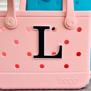 May include: A pale pink tote bag featuring a black letter "L" and red dot details. The bag has two handles and the brand name "bogg" is printed on the base. A turquoise bag is visible in the background.