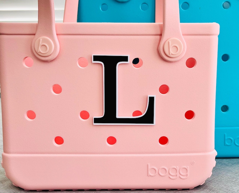 May include: A pale pink tote bag featuring a black letter "L" and red dot details. The bag has two handles and the brand name "bogg" is printed on the base. A turquoise bag is visible in the background.