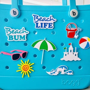 May include: A turquoise tote bag with beach-themed stickers. Stickers include the words "Beach Life" and "Beach Bum", pink sunglasses, a green and white umbrella, a red bucket and spade, a beach ball, a sandcastle, a sun, and a wave.