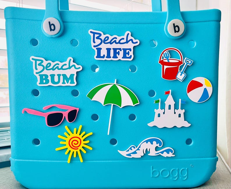 May include: A turquoise tote bag with beach-themed stickers. Stickers include the words "Beach Life" and "Beach Bum", pink sunglasses, a green and white umbrella, a red bucket and spade, a beach ball, a sandcastle, a sun, and a wave.