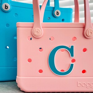 May include: Two tote bags, one light blue and one pink, with matching handles and the letter "C" on the pink bag. The bags have a perforated design with small holes and the brand name "bogg" is visible on the bags.