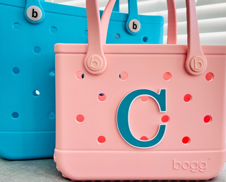 May include: Two tote bags, one light blue and one pink, with matching handles and the letter "C" on the pink bag. The bags have a perforated design with small holes and the brand name "bogg" is visible on the bags.