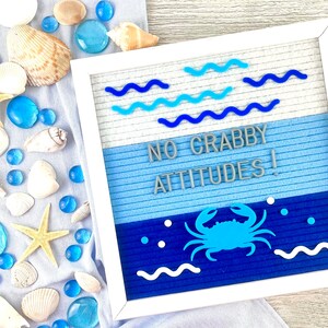 May include: A square white felt letter board featuring the text NO GRABBY ATTITUDES! in gray letters. The board has a blue gradient background with blue wavy lines, a blue crab icon, and white dots. It is surrounded by seashells and blue glass gems.