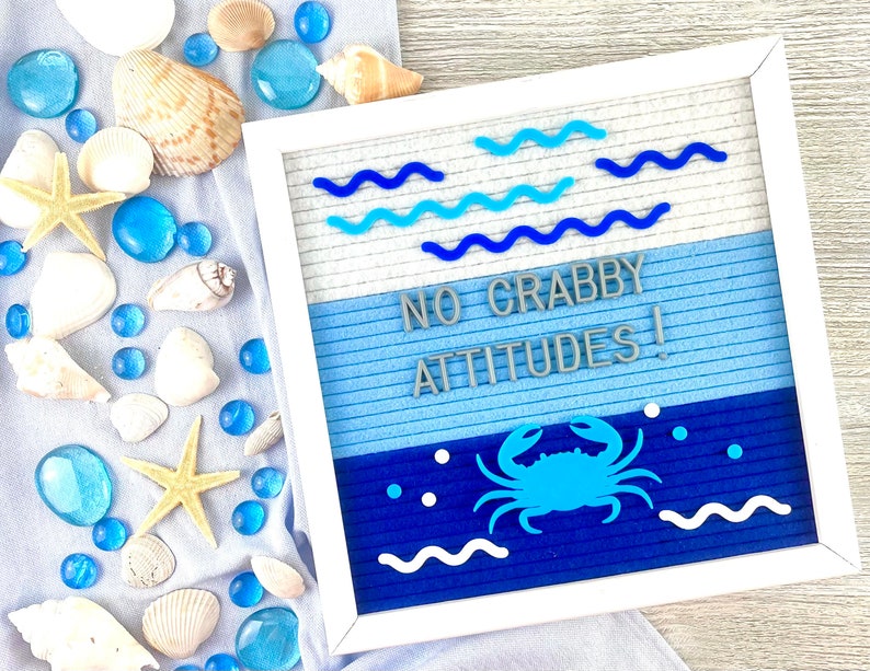 May include: A square white felt letter board featuring the text NO GRABBY ATTITUDES! in gray letters. The board has a blue gradient background with blue wavy lines, a blue crab icon, and white dots. It is surrounded by seashells and blue glass gems.