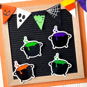 May include: A black felt letter board with a wooden frame featuring a felt Halloween bunting banner at the top and four black cauldron cutouts with bubbling liquid in purple, orange, and green colors attached to the board surface.