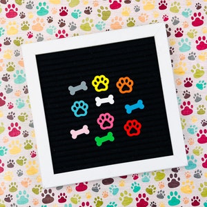 May include: A white framed black felt letter board with colorful paw print and bone-shaped magnets. The magnets are in various colors, including yellow, orange, blue, pink, and green. The background is a paw print pattern.