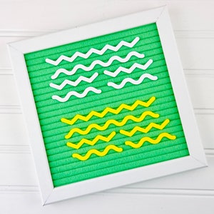May include: A square felt letter board with a bright green background and a white frame. The board features several rows of white and yellow plastic wavy and zigzag lines arranged in horizontal patterns across the grooved felt surface.