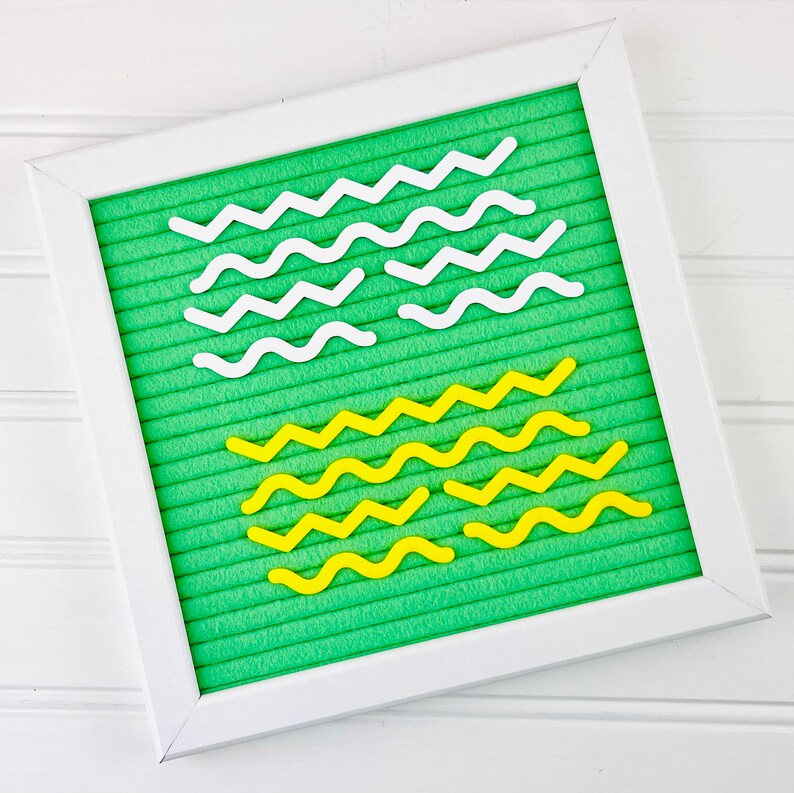 May include: A square felt letter board with a bright green background and a white frame. The board features several rows of white and yellow plastic wavy and zigzag lines arranged in horizontal patterns across the grooved felt surface.