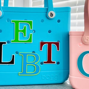 May include: Two tote bags, one large and one small, in turquoise and pink. The turquoise bag has the letters L, E, T, and B in various colours. The pink bag has the letter C. Both bags have handles and the brand logo.