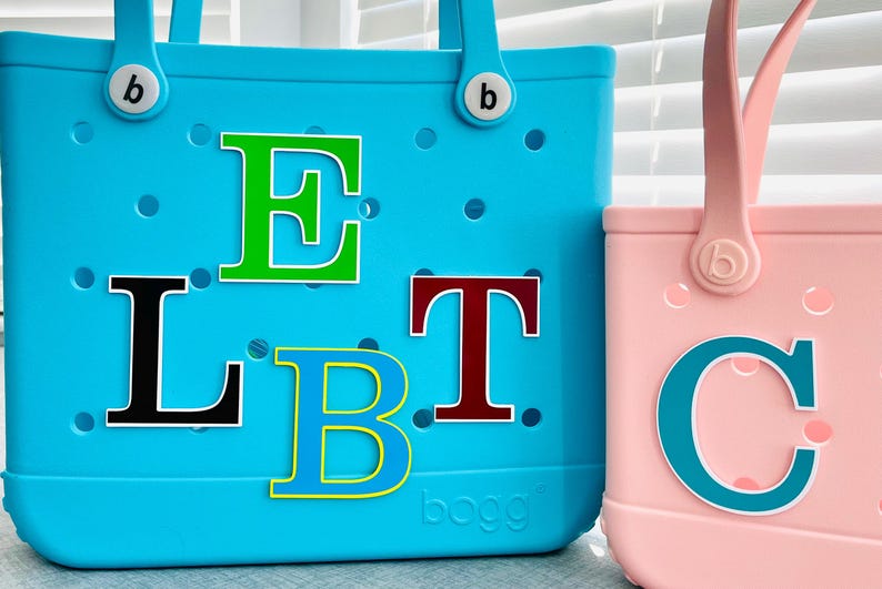 May include: Two tote bags, one large and one small, in turquoise and pink. The turquoise bag has the letters L, E, T, and B in various colours. The pink bag has the letter C. Both bags have handles and the brand logo.