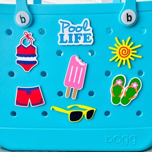 May include: A turquoise tote bag adorned with summer-themed stickers. Stickers include a red and blue swimsuit, red shorts, a pink ice lolly, yellow sunglasses, green flip-flops, a yellow sun, and the words "Pool Life."