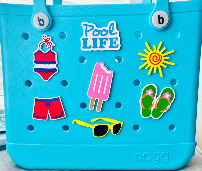 May include: A turquoise tote bag adorned with summer-themed stickers. Stickers include a red and blue swimsuit, red shorts, a pink ice lolly, yellow sunglasses, green flip-flops, a yellow sun, and the words "Pool Life."