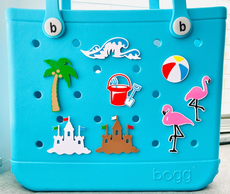 K&ouml;nnte beinhalten: Eine t&uuml;rkisfarbene Tragetasche mit Strandmotiven. Die Motive umfassen eine Palme, Wellen, einen Strandball, einen Sandeimer, eine Schaufel, zwei Flamingos und zwei Sandburgen. Auf der Tasche steht das Wort "bogg".