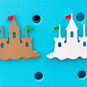 K&ouml;nnte beinhalten: Zwei sandburgf&ouml;rmige Anh&auml;nger mit bunten Flaggen. Einer ist braun, der andere wei&szlig;, beide mit roten, gr&uuml;nen und blauen Flaggen. Auf einem hellblauen Hintergrund mit kreisf&ouml;rmigen Aussparungen.