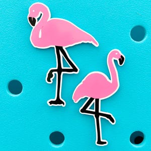 K&ouml;nnte beinhalten: Zwei rosa Flamingo-Aufkleber mit schwarzen Beinen und Schn&auml;beln werden vor einem hellt&uuml;rkisen Hintergrund gezeigt. Die Flamingos sind in verschiedenen Posen, einer steht und der andere hat das Bein gehoben. Die Aufkleber haben eine wei&szlig;e Umrandung.