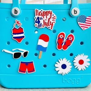 May include: A turquoise tote bag adorned with various stickers. Stickers include a swimsuit, sunglasses, a popsicle, flip-flops, daisies, and a heart-shaped American flag. The text "Happy 4th of July" is also visible.