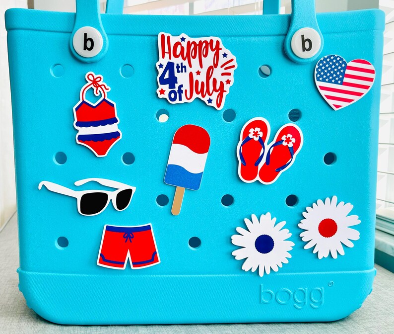 May include: A turquoise tote bag adorned with various stickers. Stickers include a swimsuit, sunglasses, a popsicle, flip-flops, daisies, and a heart-shaped American flag. The text "Happy 4th of July" is also visible.