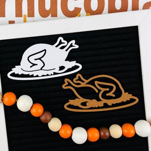 May include: A black felt letter board with a white frame features two turkey-shaped cutouts, one white and one brown. A string of wooden beads in orange, white, and brown colors is draped across the board. The background shows partial text.