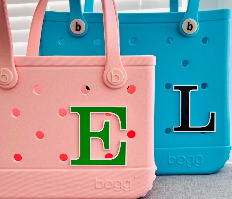 May include: Two brightly coloured tote bags. One is pink with a green letter "E" and red polka dots. The other is blue with a black letter "L". Both bags have handles and the brand name "bogg" on the bottom.