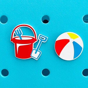 K&ouml;nnte beinhalten: Ein roter Eimer mit einer blauen Gabel, eine blaue Schaufel und ein Strandball mit roten, gelben, blauen und wei&szlig;en Streifen werden auf einem t&uuml;rkisfarbenen Hintergrund angezeigt. Die Artikel sind wahrscheinlich Strandaccessoires.