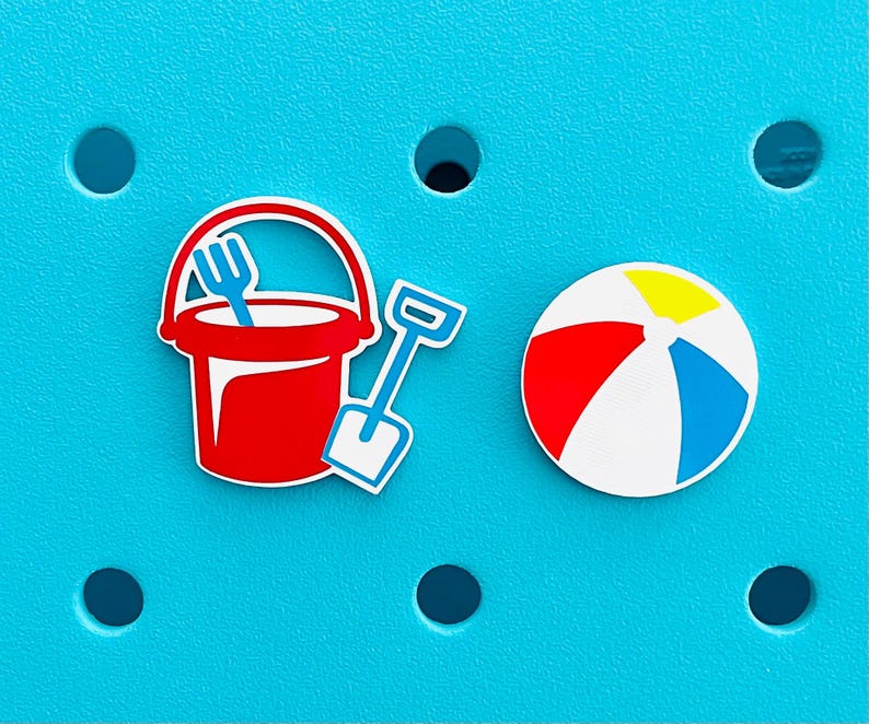K&ouml;nnte beinhalten: Ein roter Eimer mit einer blauen Gabel, eine blaue Schaufel und ein Strandball mit roten, gelben, blauen und wei&szlig;en Streifen werden auf einem t&uuml;rkisfarbenen Hintergrund angezeigt. Die Artikel sind wahrscheinlich Strandaccessoires.