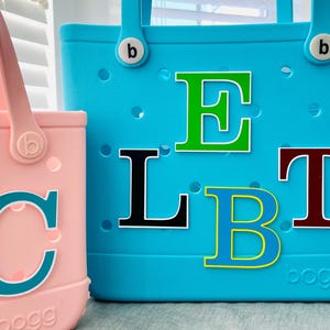 May include: Two brightly coloured tote bags. One is pink with a teal letter "C". The other is turquoise with the letters "ELBT" in various colours. Both bags have handles and the brand name "bogg" is visible.