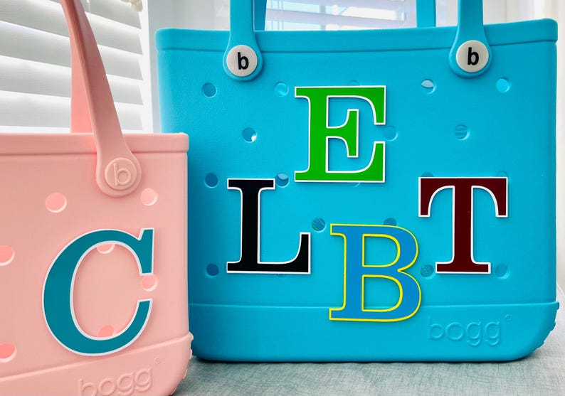 May include: Two brightly coloured tote bags. One is pink with a teal letter "C". The other is turquoise with the letters "ELBT" in various colours. Both bags have handles and the brand name "bogg" is visible.