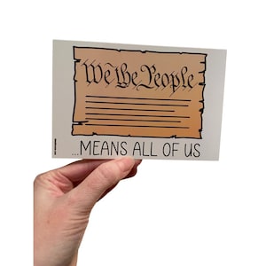 May include: A white sticker with a cartoon drawing of the beginning of the US Constitution, "We the People..." with a brown parchment background. The text below the drawing says "...means all of us".
