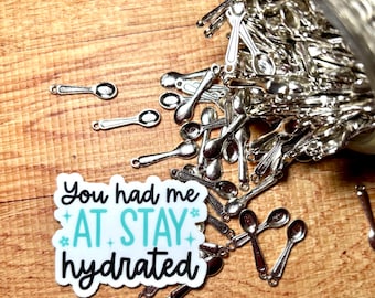 May include: A collection of small silver-colored metal spoon charms spilling from a glass jar next to a white rectangular sticker with the black and teal text: You had me AT STAY hydrated, decorated with small star and flower icons.