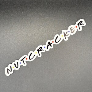 May include: A white sticker with the word "NUTCRACKER" in black, with each letter having a different color dot. The sticker is on a dark gray surface. The design is reminiscent of a popular television show.