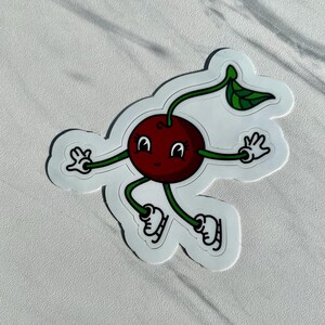 May include: A sticker featuring a cartoon cherry with a smiling face, arms, and legs. The cherry is red with a green stem and leaf. The cherry is wearing ice skates and has its arms outstretched.