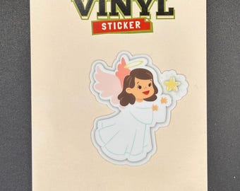 May include: A vinyl sticker featuring a cartoon angel with pink wings, a halo, and a white dress, holding a star. The sticker is on a beige background with the words "VINYL STICKER" in green and red.
