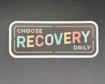 May include: A rectangular sticker with rounded corners. The sticker has a black background with the words "CHOOSE RECOVERY DAILY" in colorful block letters. The text is outlined in white and framed by a thin black border.