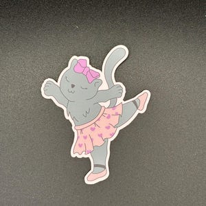 May include: A gray cat sticker in a ballet pose. The cat wears a pink bow, a pink tutu with heart shapes, and pink ballet slippers. The cat's arms are outstretched, and one leg is extended.