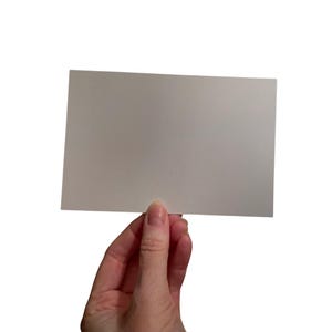 Puede incluir: Una tarjeta rectangular blanquecina sostenida por una mano sobre un fondo blanco. La tarjeta es lisa y parece estar en blanco, lo que sugiere que podr&iacute;a ser una postal o una tarjeta de notas.