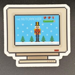 May include: A sticker featuring a pixelated computer screen displaying a winter scene with the text "THE NUTCRACKER." The screen shows a nutcracker soldier, snowflakes, and Christmas trees. The sticker has a white border.