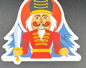 May include: A sticker featuring a cartoon nutcracker soldier in a red uniform with gold accents, a black hat, and a white beard. The background includes a red circle and blue stylized trees, with a silver sword in hand.